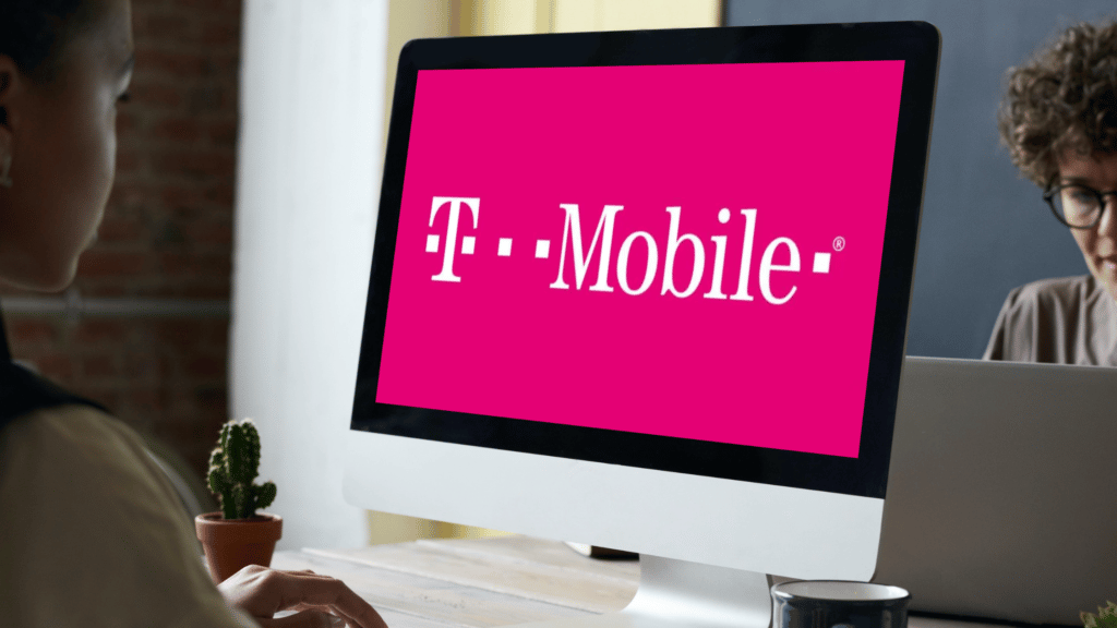 mulher sentada em frente a tela de um computador, onde é possível ver o logo da T-Mobile escrito em branco sobre o fundo rosa. Atrás do computador é possível ver uma parte do rosto de uma pessoa branca usando óculos