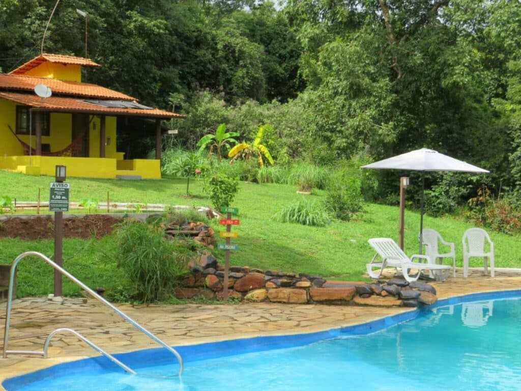 Chalé em Pirenópolis com piscina, cadeiras de praia e as acomodações ao fundo, rodeado por vegetação.