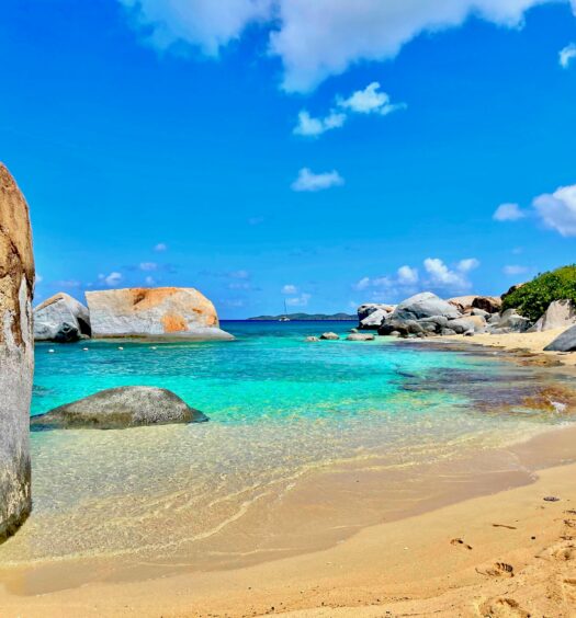 Praia com água cristalina, em um dia de céu aberto e de sol, com pedras grandes espalhadas, além de ter areia fofa e bege. Imagem para ilustrar o post chip celular Ilhas Virgens Britânicas