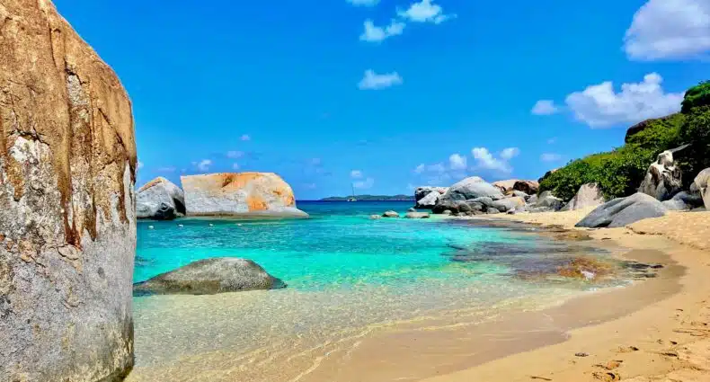 Praia com água cristalina, em um dia de céu aberto e de sol, com pedras grandes espalhadas, além de ter areia fofa e bege. Imagem para ilustrar o post chip celular Ilhas Virgens Britânicas