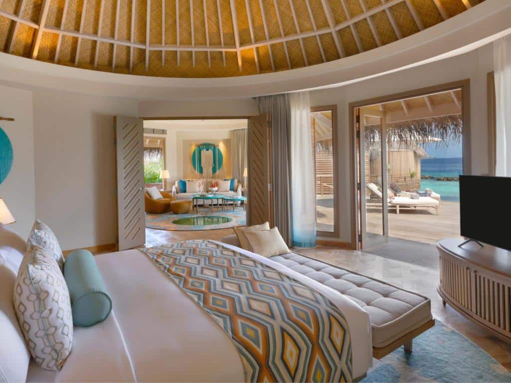 Interior de uma acomodação do The Nautilus Maldives, com cama de casal, móvel com TV e sofá à frente. Há uma varanda com deck de madeira e vista para o mar cristalino e, no lado esquerdo, há uma sala de estar super bem decorada