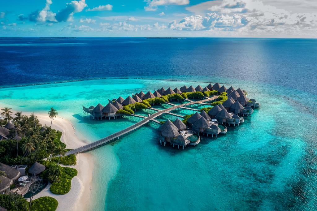Vista aérea de bangalôs espalhados no mar do The Nautilus Maldives, com um mar azulado num tom claro mais próximo da costa e num tom mais escuro nas redondezas. Há também parte de uma ilha com natureza do hotel. Imagem para ilustrar os hotéis em Maldivas