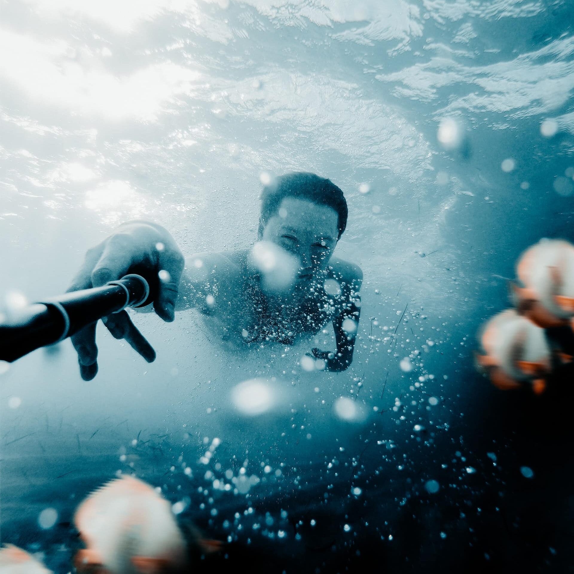 Homem no fundo do mar segurando um extensor de camera, com bolhas da água em sua volta.