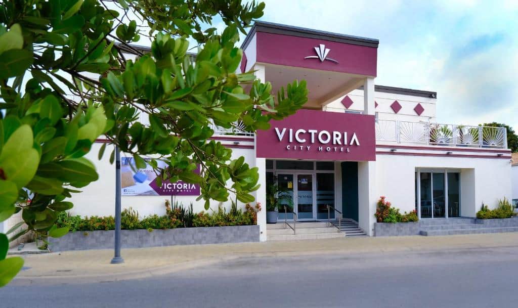 frente do Victoria City Hotel em Aruba com alguns galhos e folhas aparecendo no canto esquerdo da imagem. O hotel é branco com detalhes em vinho.