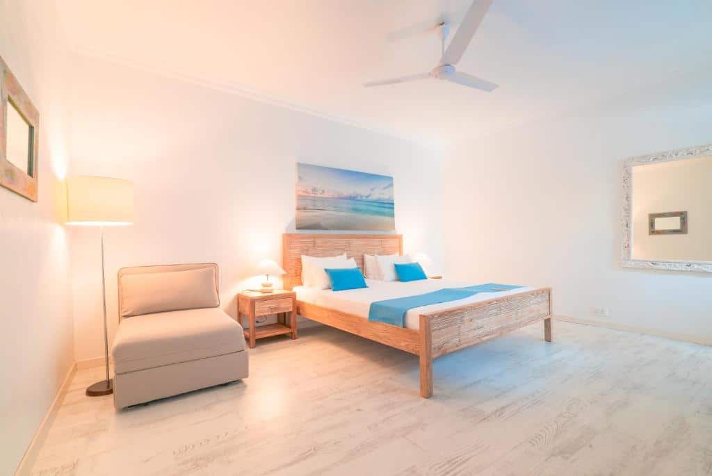 Quarto deluxe da Villa Rosa Maldives, de 32 m², com uma cama de casal e poltrona ao lado. Há bastante espaço em volta da cama, um ventilador de teto e um espelho na parede direita