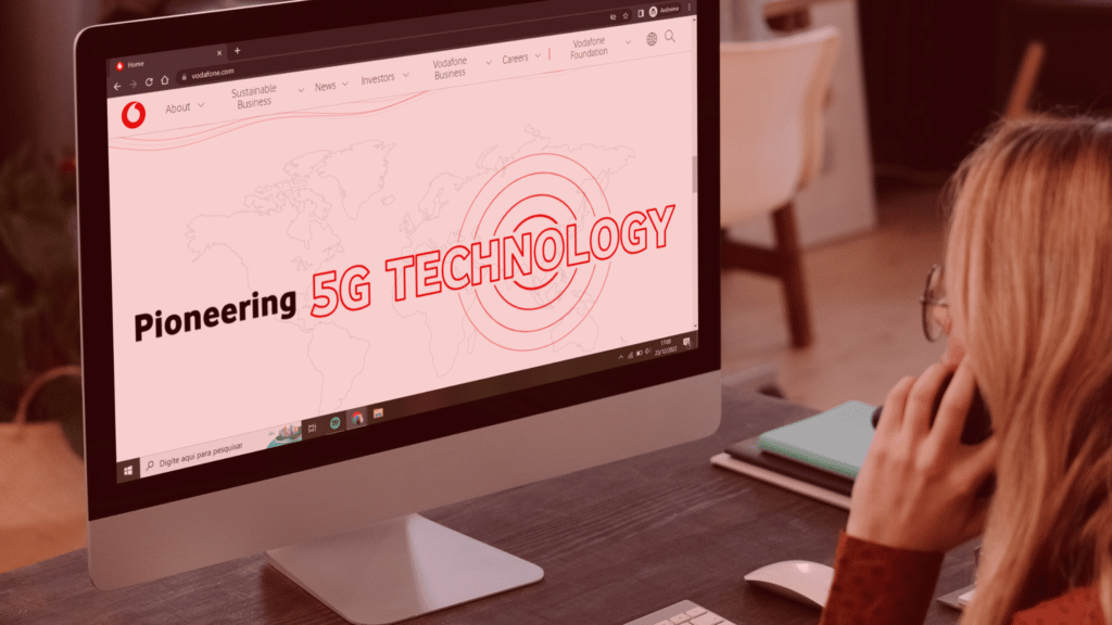 mulher loira, com cabelo liso, está sentada em frente a um grande monitor de computador com a tela mostrando o site da empresa Vodafone. Na tela é possível ler "Pioneering 5G Technology" - que significa "Pioneiro em tecnologia 5g".