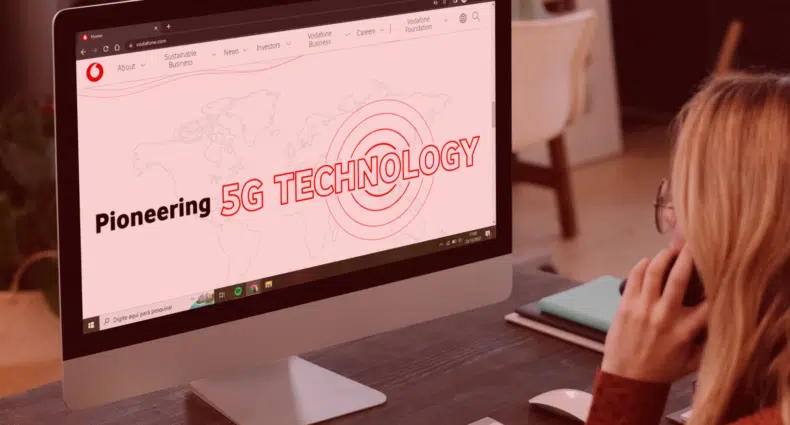 mulher loira, com cabelo liso, está sentada em frente a um grande monitor de computador com a tela mostrando o site da empresa Vodafone. Na tela é possível ler "Pioneering 5G Technology" - que significa "Pioneiro em tecnologia 5g".