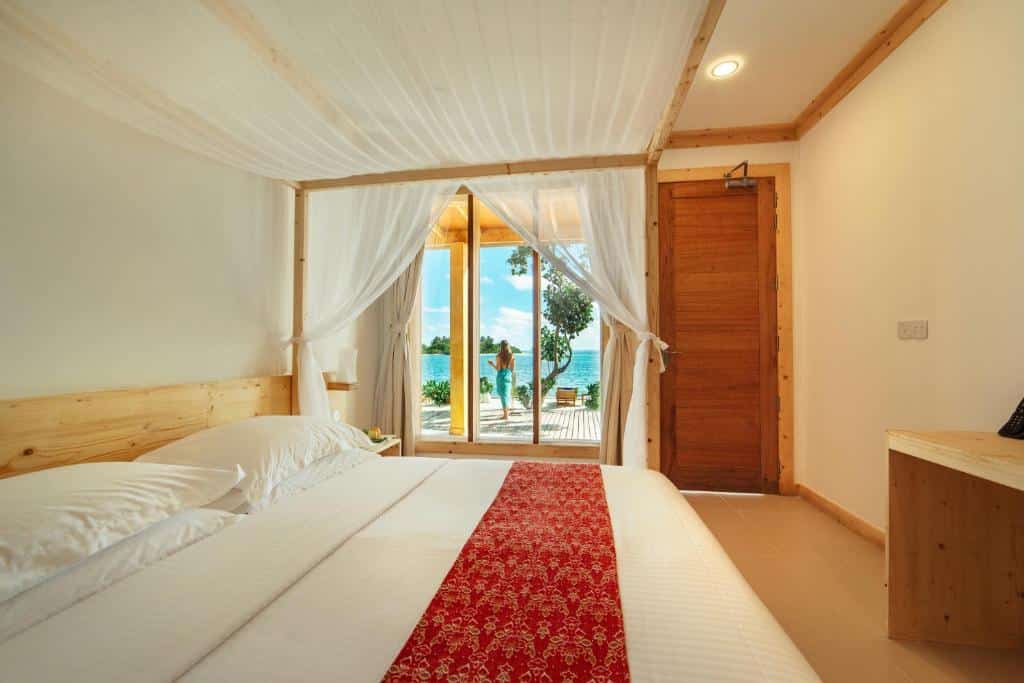 Quarto duplo, de 24 m², do Wave Sound, um dos hotéis em Maldivas. Nele, há uma cama de casal, uma cortina branca e uma porta de vidro mostrando um moça em um deck à beira-mar, com uma praia cristalina