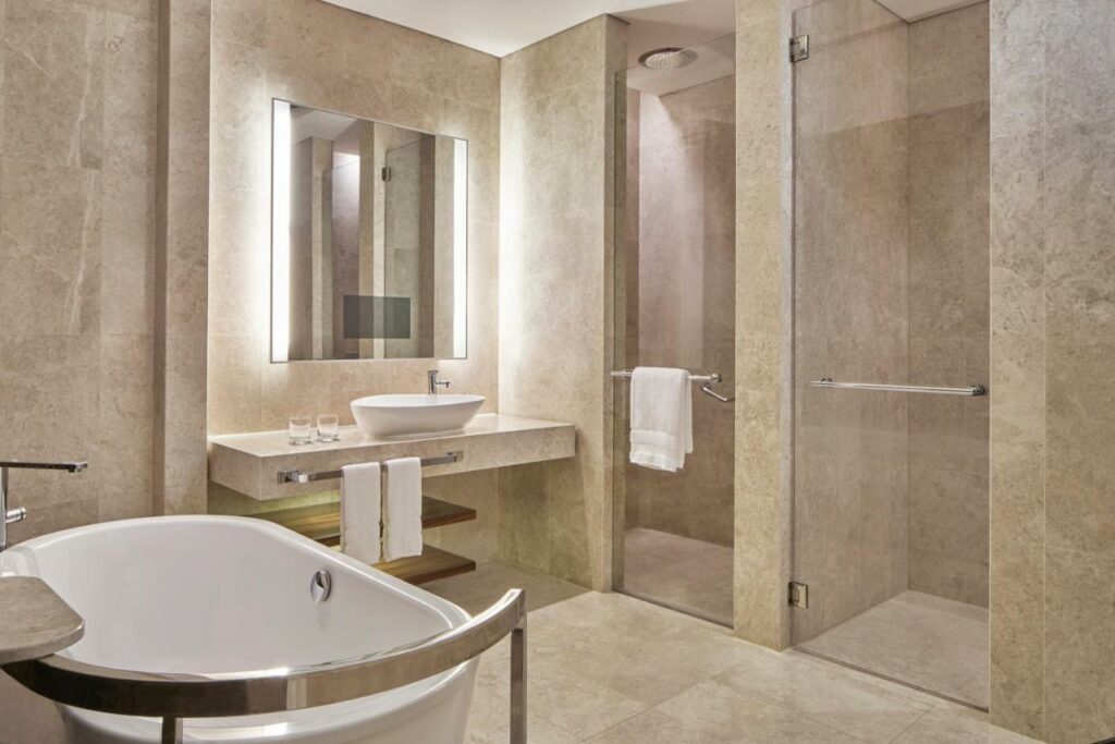 Banheiro de um quarto do hotel The Westin Singapore. O cômodo é todo de mármore bege e tem uma banheira encostada na parede da esquerda. Duas portas de vidro tem chuveiro e privada e, na parede central, a pia de mármore tem um espeljo acima e algumas toalhas no suporte abaixo. Dois copos de vidro estão do lado da pia.