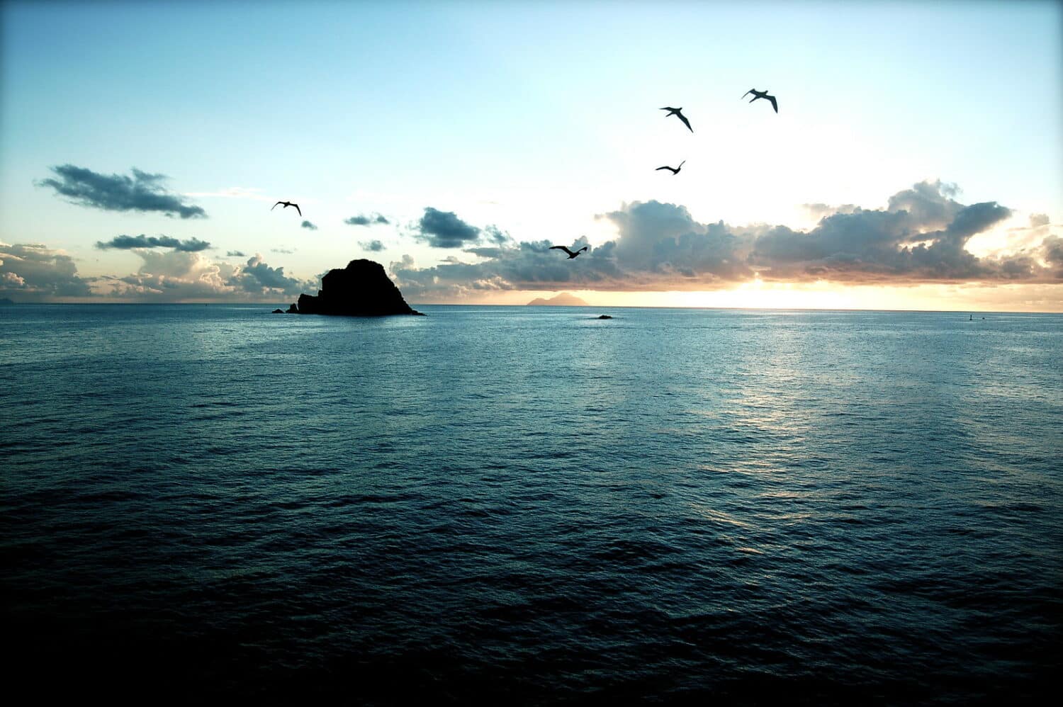 Pôr do sol na praia em St. Barth. Há uma ilhota ao loge, e pássaros voam acima. Há poucas nuvens no céu, que está azul e laranja. - Foto: Keith Robinson via Flickr