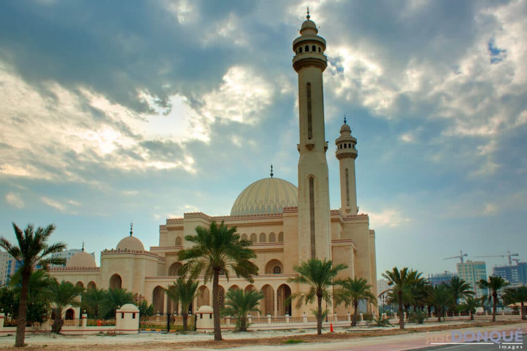 vista da lateral da Grande Mesquita Al Fateh, em Manama, com duas torres iguais de ambos os lados da mesquita, que tem uma abóbada e outros detalhes e tons de rosa claro em um bege suave da sua arquitetura trabalhada, há coqueiros na frente e o céu está azul com muitas nuvens e raios de sol, para ilustrar o post de chip celular Bahrein