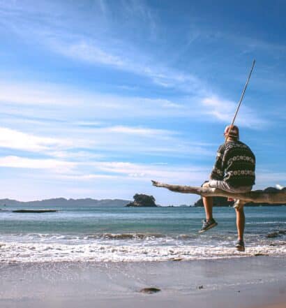 vista de uma praia com poucas ondas e águas esverdeadas e céu azul, em Cathedral Cove, na Nova Zelândia, com uma pessoa sentada em um tronco de árvore segurando uma vara de pesca, para ilustrar o post de chip celular Oceania