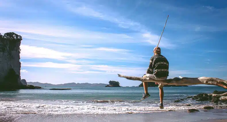 vista de uma praia com poucas ondas e águas esverdeadas e céu azul, em Cathedral Cove, na Nova Zelândia, com uma pessoa sentada em um tronco de árvore segurando uma vara de pesca, para ilustrar o post de chip celular Oceania