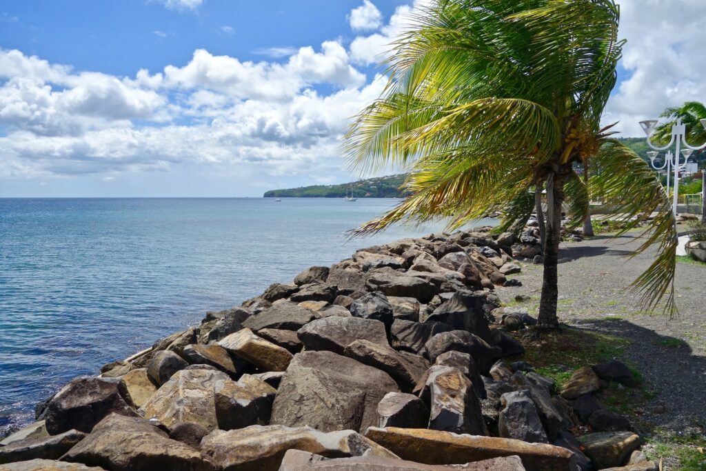 vista da praia de La Trinité, com bastante pedras que separam da praia de águas e com algumas árvores e cadeirinhas brancas ao fundo e o céu está azul com nuvens, para ilustrar o post de chip celular Martinica