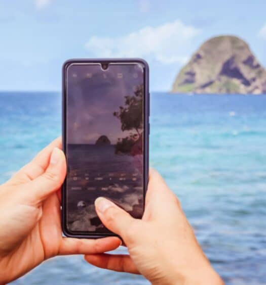 mãos brancas segurando um celular em que tira uma foto de uma formação rochosa em meio ao mar cristalino e céu azul em