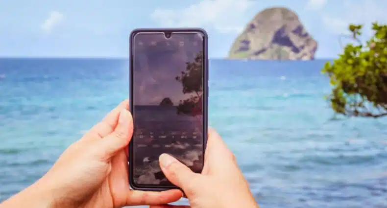 mãos brancas segurando um celular em que tira uma foto de uma formação rochosa em meio ao mar cristalino e céu azul em