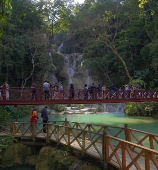 vista de várias pessoas em uma ponte vermelha em Luang Prabang, Laos, com uma cachoeira ao fundo e águas plácidas, com vegetação ao redor, para ilustrar o post de chip celular Ásia