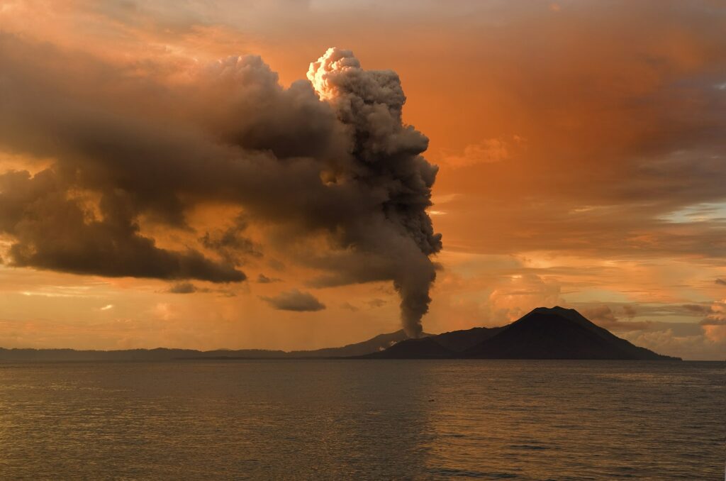 vista do Vulcão Tavurvur, na Nova Zelândia, em tons de amarelo e laranja com águas calmas e o vulcão ao longe expelindo fumaça que dá acesso às águas termais, para ilustrar o post de chip celular Oceania