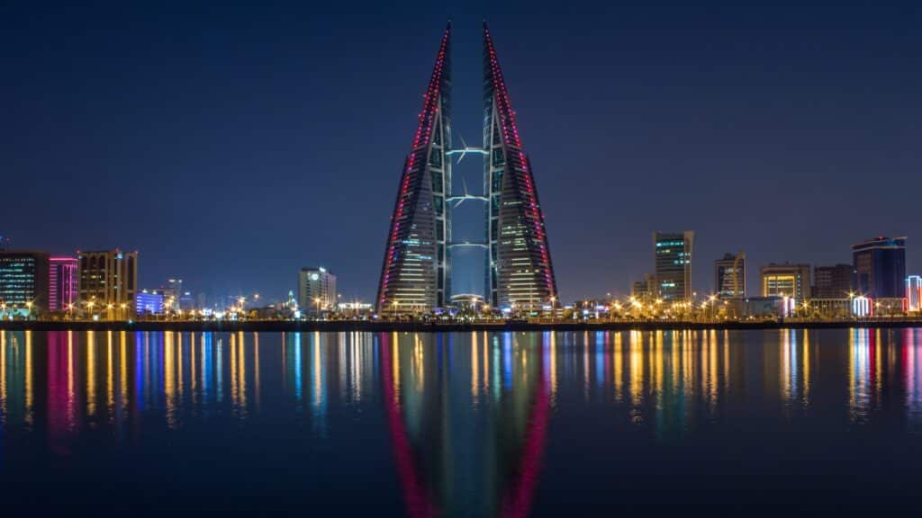 vista noturna da World Trade Center, em Manama, duas torres muito iluminadas em vermelho e azul formando um arco reto que não se encontra no topo, há mais luzes abaixo dela e tudo está refletido na água abaixo, para ilustrar o post de chip celular Bahrein