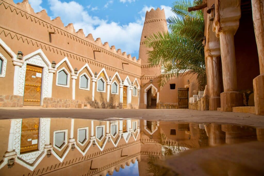 Construção marrom e branca com uma palmeira à frente para ilustrar o post sobre chip de celular para a Arábia Saudita. O céu azul e o prédio são refletidos em uma poça de água no centro. - Foto: Abdulrhman Alkhnaifer via Unsplash