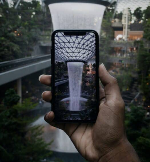 Mão segurando um celular com a câmera tirando foto de uma cascata alta com escultura de vidro acima, no Aeroporto de Singapura, para ilustrar o post de chip celular Singapura