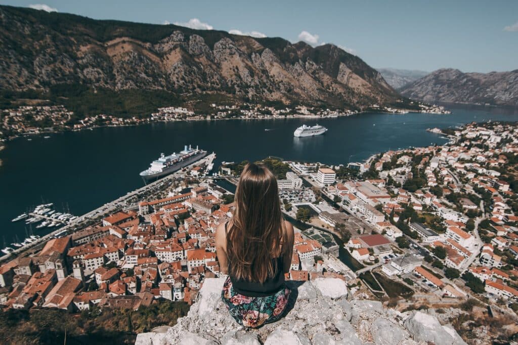 Um moça sentada em um rocha que fica no alto de uma cidade com vista para o mar, há muitas embarcações na água e algumas montanhas mais ao fundo