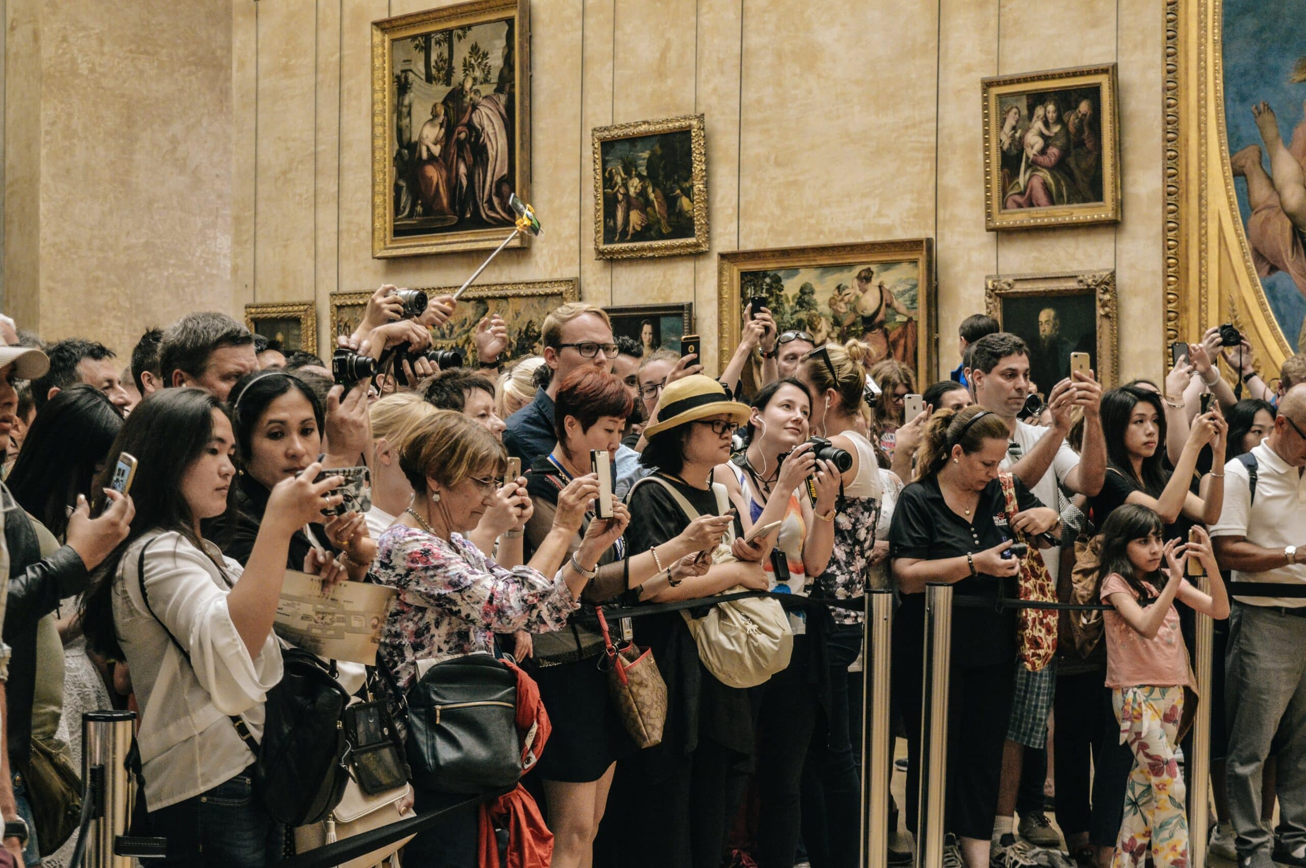 dezenas de pessoas amontadas perto da faixa de contenção em frente ao quadro de Monalisa - que não aparece na imagem - dentro do Museu do Louvre, em Paris. Ao fundo é possível ver outras obras de arte pintadas nos quadros pendurados à parede.