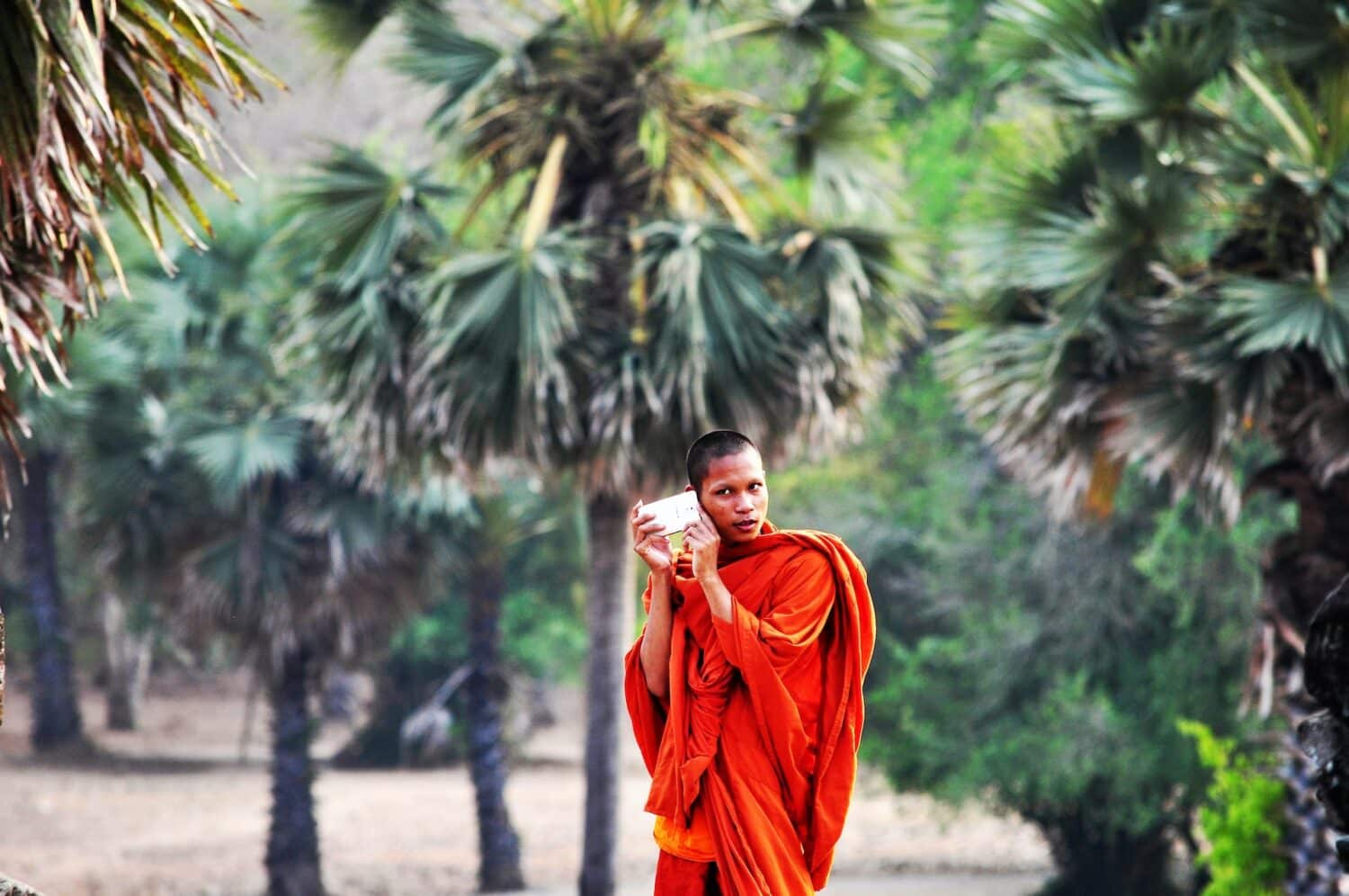 Homem monge, vestido de roupa típica laranja, segurando o celular com as duas mãos no ouvido direito, com bastante natureza atrás. Imagem para representar o post chip de celular Camboja