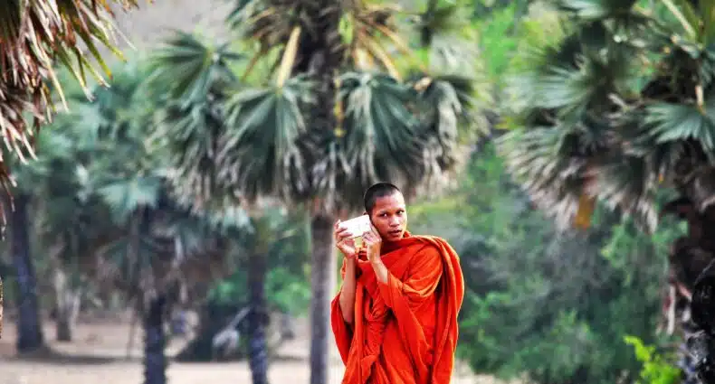 Homem monge, vestido de roupa típica laranja, segurando o celular com as duas mãos no ouvido direito, com bastante natureza atrás. Imagem para representar o post chip de celular Camboja