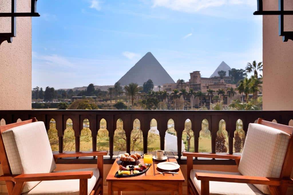 Área de café do Marriott Mena House com duas cadeiras de madeira com estofado branco com mesa no centro e ao fundo vista para as pirâmides. Representa Hotéis no Cairo.