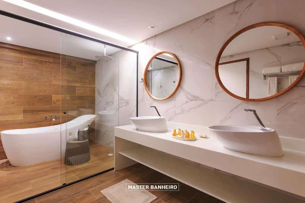 Banheiro amplo do Armação Resort Porto de Galinhas com uma banheira dentro de um box amplo, sendo possível tomar banho em qualquer uma das opções, com uma pia com duas cubas, dois espelhos redondos, para representar resorts em Muro Alto
