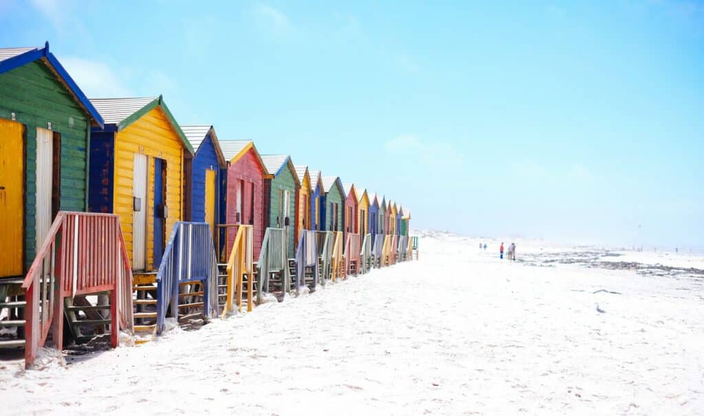Uma praia de areia branca com diversas casinhas de madeira coloridas com algumas pessoas caminhando pela região
