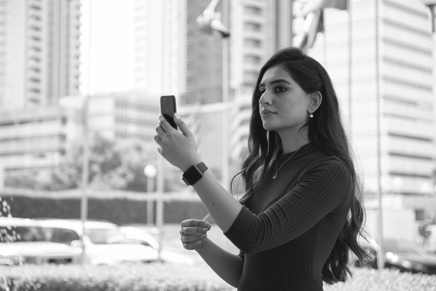 mulher segurando um celular olhando para tela, ela tem traços árabes e a foto está em preto e branco, há prédios atrás e o que parece ser uma via movimentada, para ilustrar o post de chip celular Bahrein