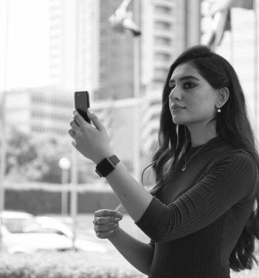 mulher segurando um celular olhando para tela, ela tem traços árabes e a foto está em preto e branco, há prédios atrás e o que parece ser uma via movimentada, para ilustrar o post de chip celular Bahrein