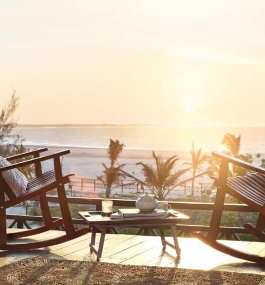 Varanda da Baía das Caraúbas, uma das pousadas em Camocim, com duas cadeiras de balanço de madeira, uma mesinha no centro com um bule de chá, e vista para o mar com o pôr do sol refletindo