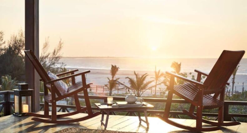 Varanda da Baía das Caraúbas, uma das pousadas em Camocim, com duas cadeiras de balanço de madeira, uma mesinha no centro com um bule de chá, e vista para o mar com o pôr do sol refletindo