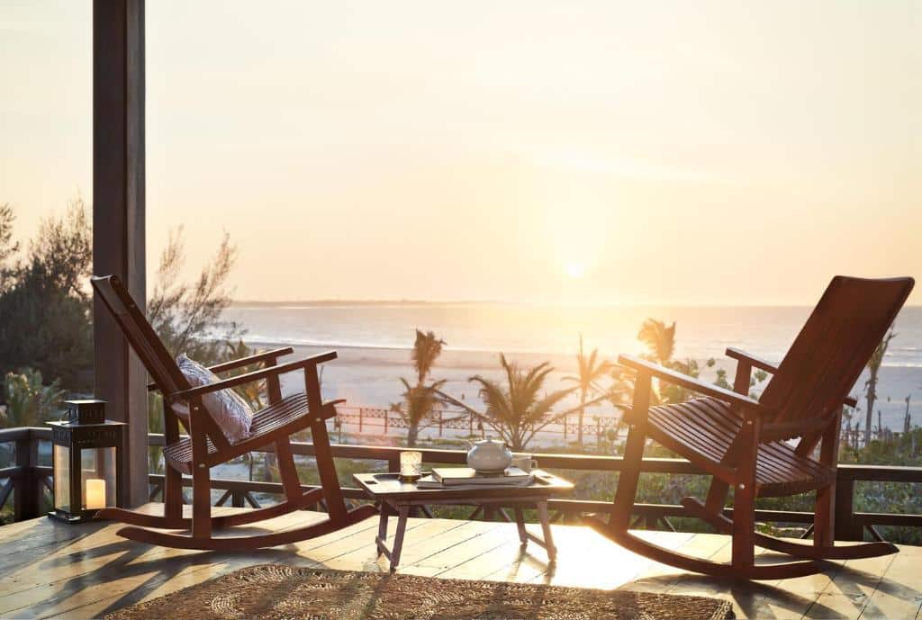 Varanda da Baía das Caraúbas, uma das pousadas em Camocim, com duas cadeiras de balanço de madeira, uma mesinha no centro com um bule de chá, e vista para o mar com o pôr do sol refletindo