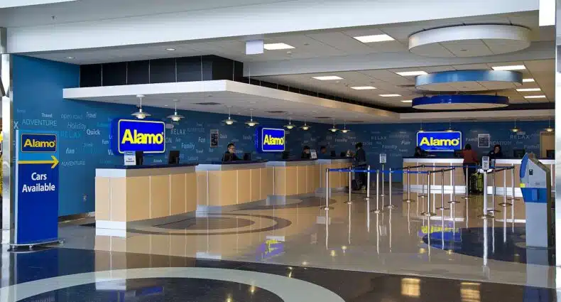 Loja da Alamo Rent a Car, com balcões quase vazios, boa iluminação, atendentes em computadores e parede com palavras em diversas línguas