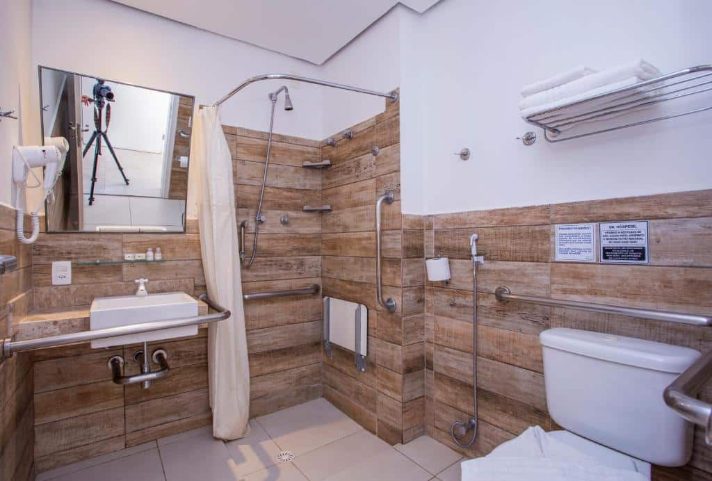 Vista do banheiro com mobilidade reduzida do Ciribaí Praia Hotel com chuveiro com barras de segurança, pia baixa com barras e vaso sanitário com barra de segurança.