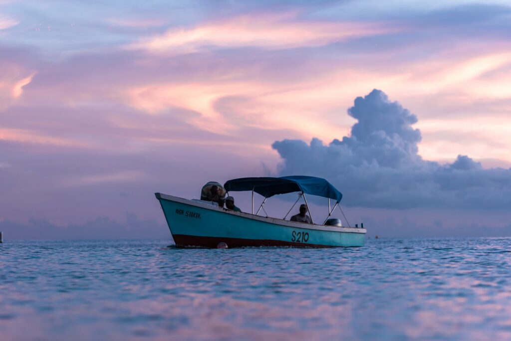 Barco no mar em Barbados, ao entardecer, com parte da água refletindo as cores arroxeadas do céu de crepúsculo, e uma nuvem azul pastel ao fundo