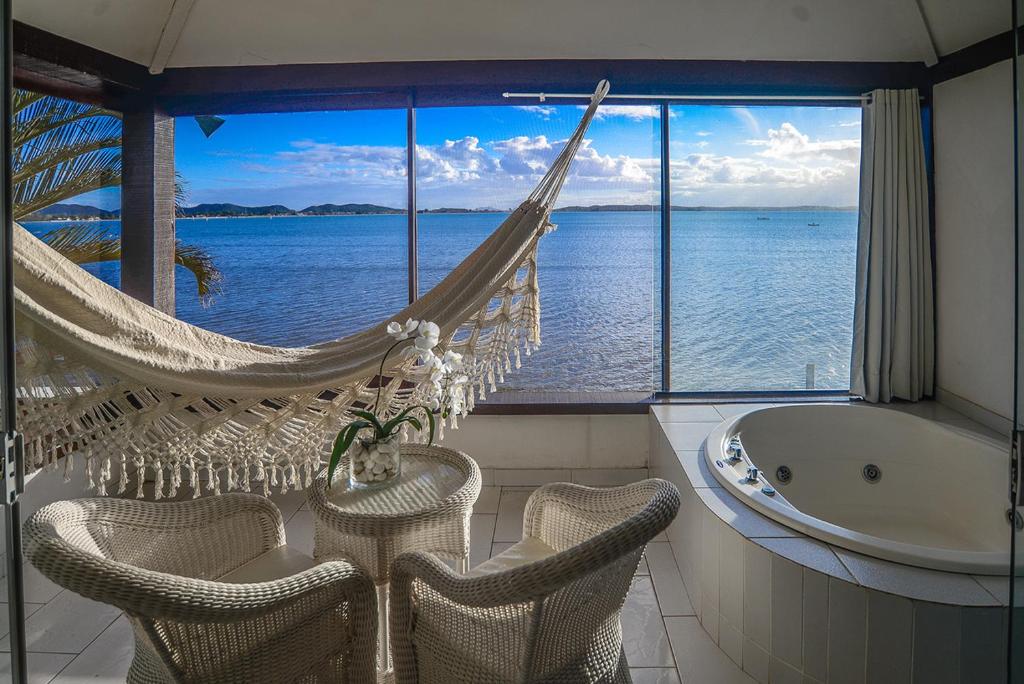 Sacada do Barracuda Eco Resort Búzios com uma janela de vidro grande dando vista para o mar, uma banheira de hidro, uma rede de descanso e duas cadeiras
