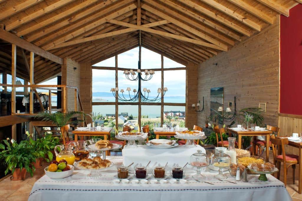 Mesa com café da manhã do Blanca Patagonia Hostería Boutique y Cabañas, sendo o ambiente rústico e com uma parede de vidro ao fundo mostrando a paisagem do lado de fora composta por um lago e montanhas