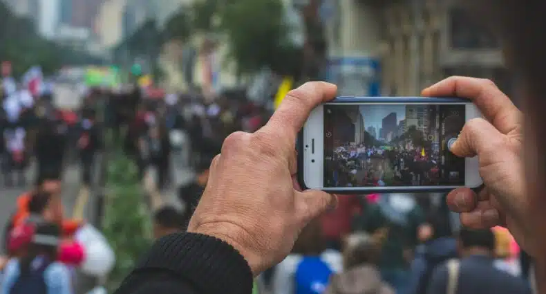 Duas mãos de um homem de idade madura segurando um celular e fotografando uma região de Bogotá, que tem várias pessoas e um rio marrom ao fundo. Imagem para ilustrar o post de chip celular Colômbia