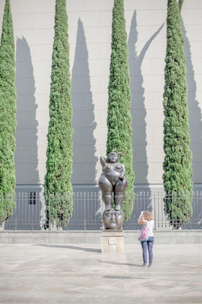 Uma mulher tirando foto de uma estátua de outra mulher em uma praça de Medelin