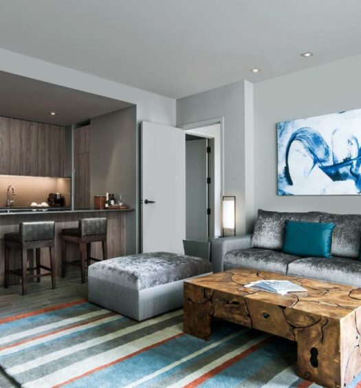 Sala de estar e cozinha compacta do East Miami, com sofá cinza com almofadas azul turquesa e do lado esquerdo cozinha, com armários de madeira. Representa hotéis com cozinha em Miami.