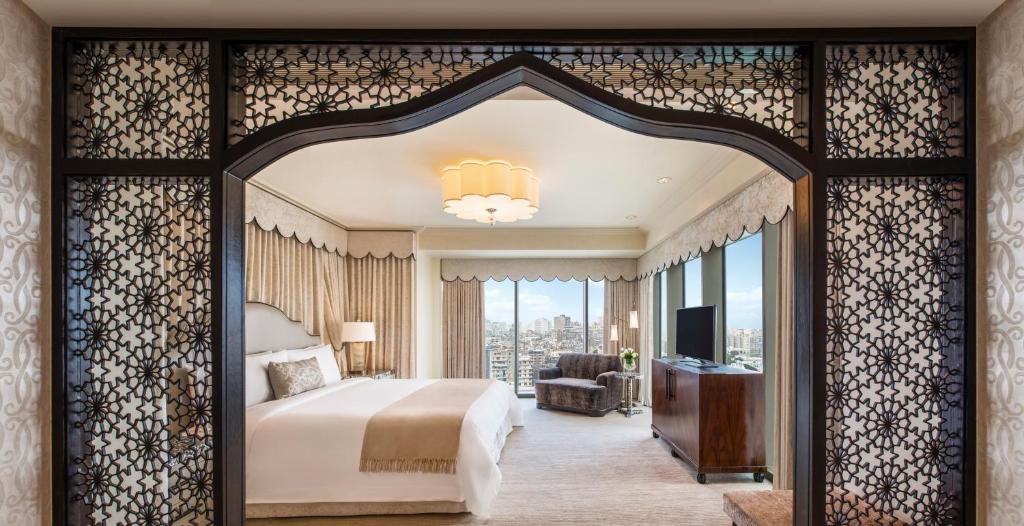 Vista do quarto do The St. Regis com cama de casal, uma cômoda ao lado esquerdo da cama com luminária e em frente a cama cômoda de madeira com TV. Representa Hotéis no Cairo.