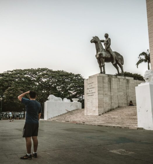 Homem vestido de blusa verde e bermuda preta, de costas, olhando o Monumento A Los Precursores. O dia está nublado e a imagem é para ilustrar o post de chip de celular Caracas