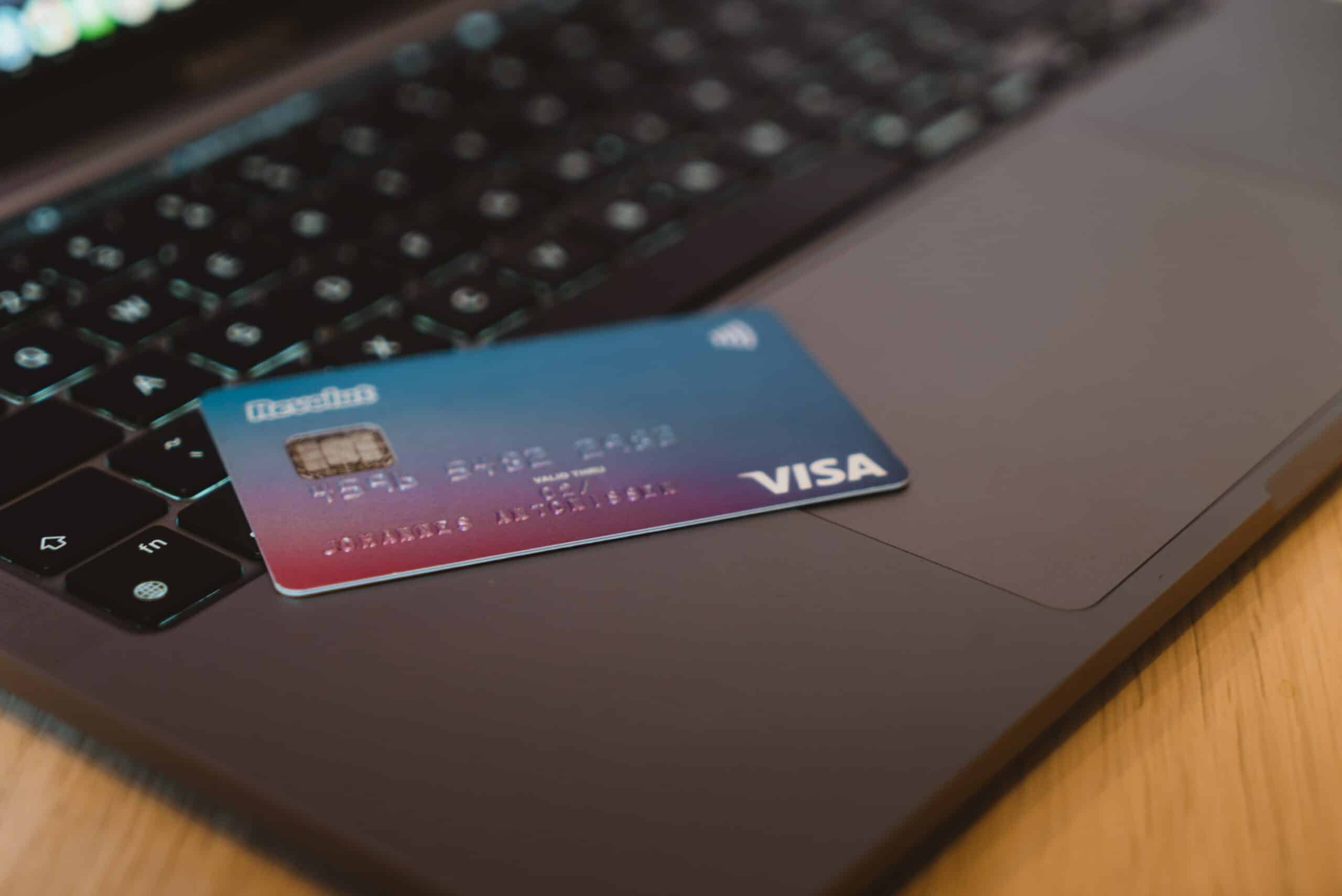 cartão de crédito azul e rosa da bandeira Visa deitado em cima de um notebook aberto. É possível ver um pequeno pedaço da tela do computador ligado