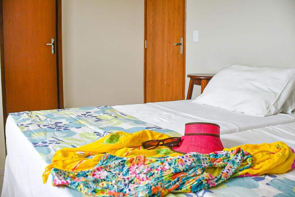 Cama de um dos quartos da Casa DasCli, com roupa de cama branca, travesseiros e lençol dobrado de estampa com tons azuis em cima, além de um óculos marrom, um chapéu rosa e roupa de praia colorida em cima da cama.