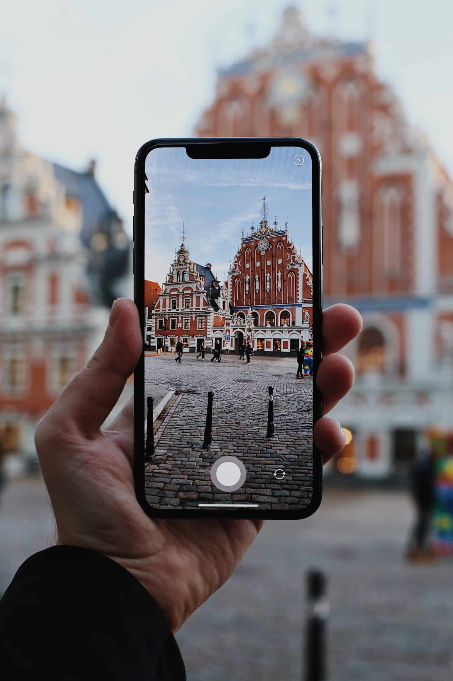 Imagem de um celular tirando foto do ponto turístico "Casa dos Cabeças Negras" na Letônia durante o dia, ilustrando post chip celular Letônia.